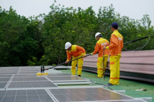 Three technicians in uniforms and hard hats cleaning rooftop solar panels.